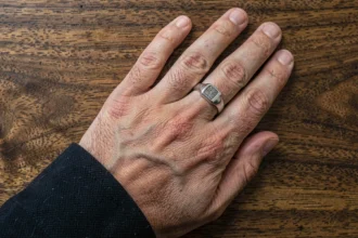 Chevalière homme en argent portée sur la main posée sur une surface en bois sombre, détail de la bague et texture de la peau visibles