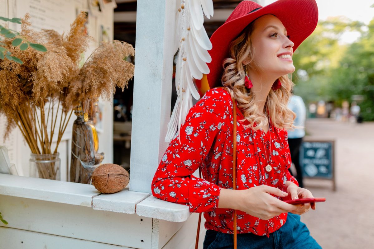 Jolie femme souriante blonde élégante en chapeau rouge de paille et tenue de mode d'été chemisier tenant à l'aide de café de téléphone intelligent